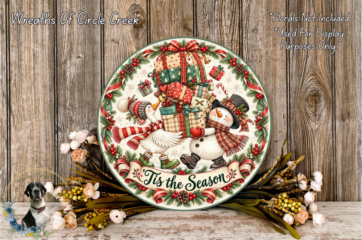A decorative Christmas wreath with a snowman, presents, and the text "It's the Season" displayed on a wooden surface.