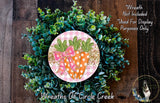 a wreath made of green foliage and two orange carrots with pink bows, placed on a wooden surface.