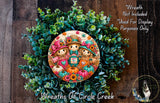 a wreath with a decorative design of three scarecrow characters, surrounded by a lush green plant. The wreath is placed on a wooden surface, and there is a small dog visible in the bottom right corner of the image.