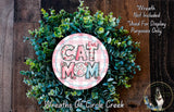 A wreath made of green leaves and a pink circular sign with the text "CAT MOM" in the center, placed on a wooden surface.