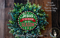 a Christmas wreath with the text &quot;Jesus is the reason for the season&quot; displayed on it. The wreath is made of green leaves and is placed on a wooden surface.