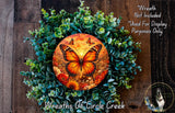 A wreath made of green foliage and flowers, with a large orange butterfly in the center. The wreath is displayed on a wooden surface, and there is a small dog in the bottom right corner.