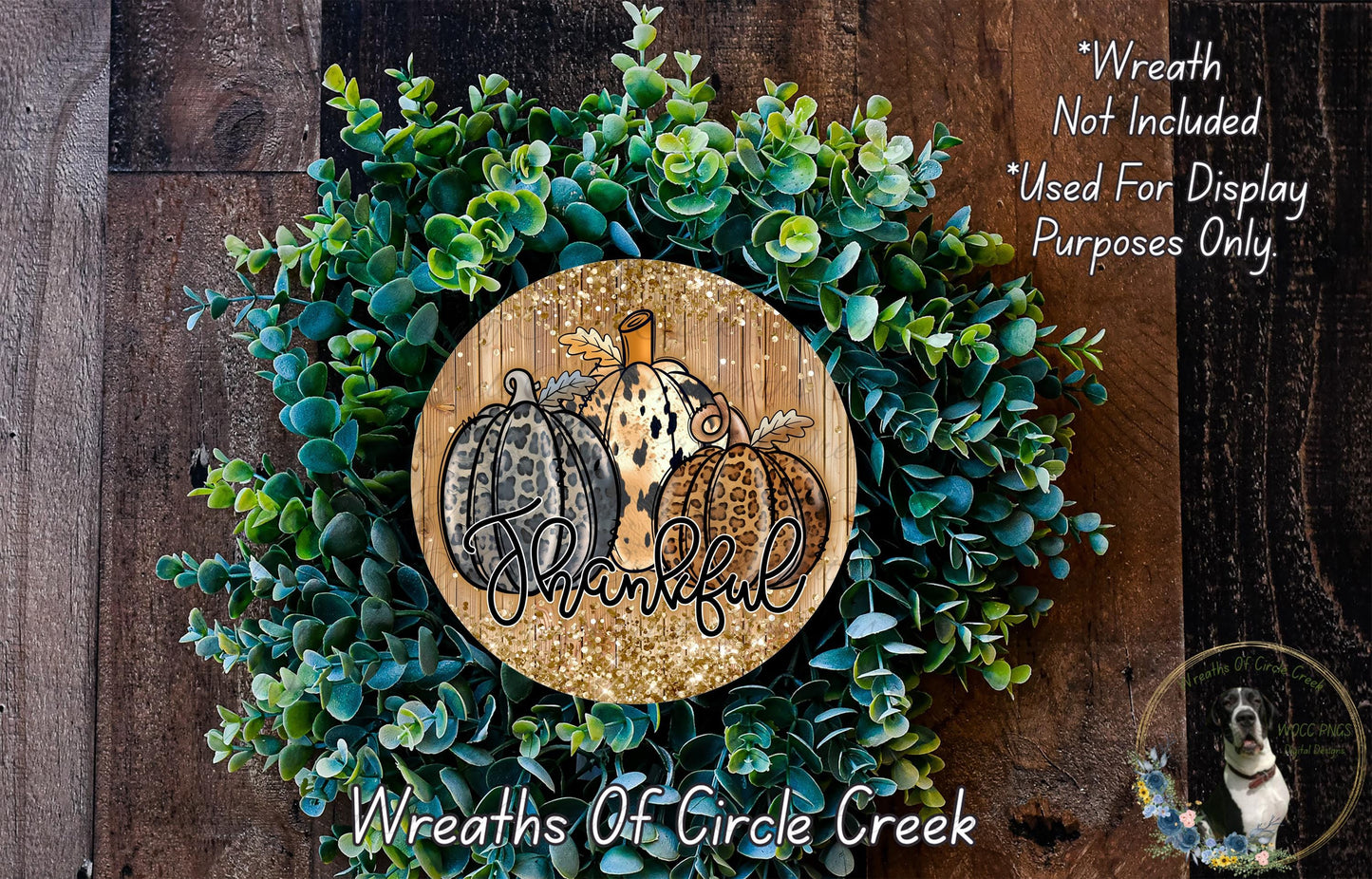 A wreath made of green leaves and three pumpkins, with the word "Thankful" written on it. The wreath is displayed on a wooden surface, and there is a small dog in the bottom right corner.