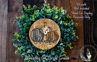 A wreath made of green leaves and three pumpkins, with the word "Thankful" written on it. The wreath is displayed on a wooden surface, and there is a small dog in the bottom right corner.