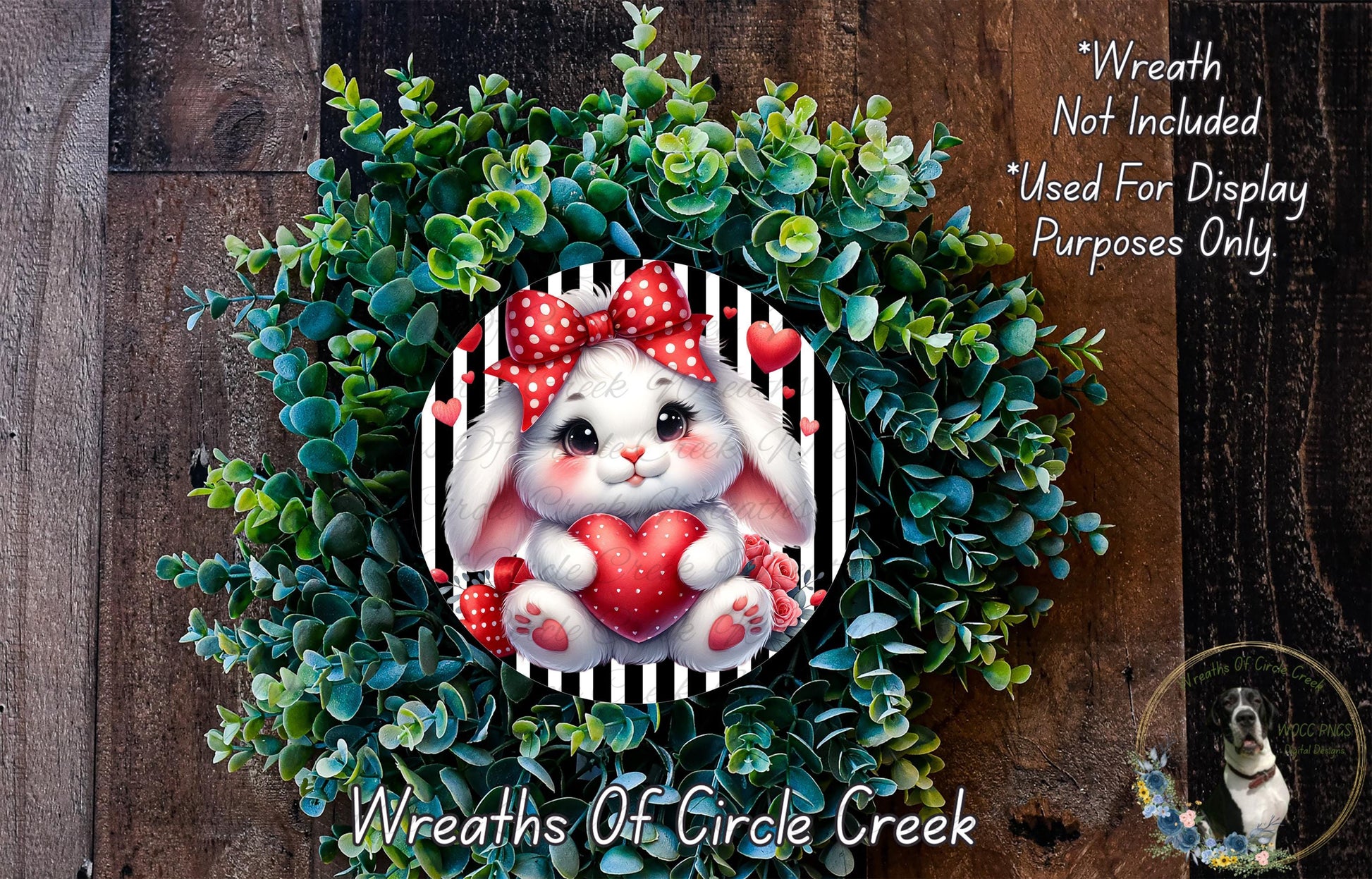 A wreath made of green leaves and a white rabbit with a red bow, holding a red heart, is displayed on a wooden surface.