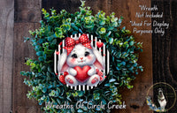 A wreath made of green leaves and a white rabbit with a red bow, holding a red heart, is displayed on a wooden surface.