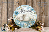 A decorative welcome sign with a sleeping lamb and flowers, placed on a wooden surface.