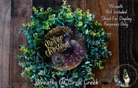 A wreath made of green and purple plants, with a gold and purple peacock feather design, and the text "Merry Christmas" written on it.