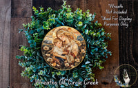 A wreath made of green leaves and flowers, with a painting of a woman holding a child in the center. The wreath is displayed on a wooden surface, and there is a text overlay that reads "Wreath Not Included * Used For Display Purposes Only".