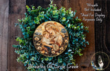 A wreath made of green leaves and flowers, with a painting of a woman holding a child in the center. The wreath is displayed on a wooden surface, and there is a text overlay that reads "Wreath Not Included * Used For Display Purposes Only".