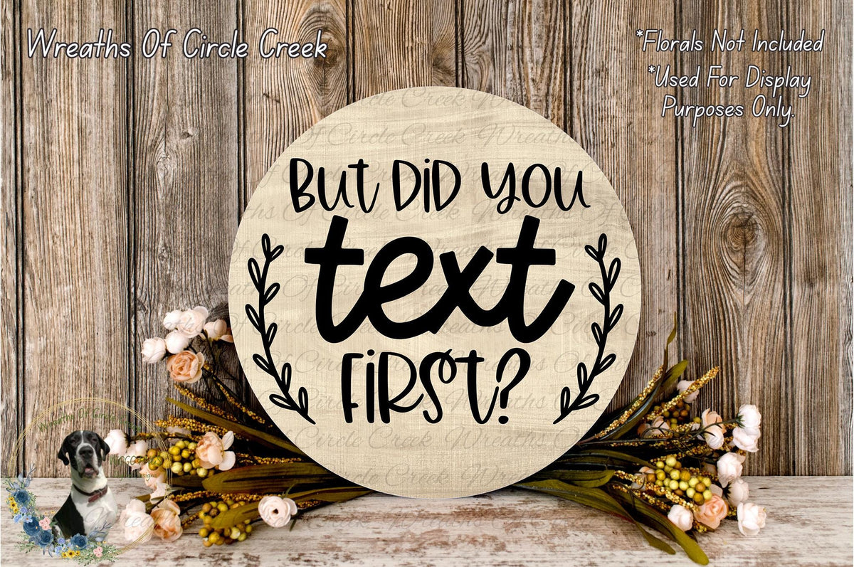 A wooden sign with the text "But Did You Text First?" surrounded by flowers and greenery, placed on a wooden surface.