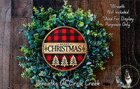 A Christmas wreath made of evergreen branches and a circular sign with a red and black plaid pattern, featuring the text "Merry Christmas" and three Christmas trees.
