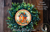 a wreath with a house and greenery, including a small orange house, on a wooden background.