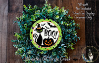 A Halloween-themed wreath made of green foliage and a black cat wearing a witch's hat, with a carved pumpkin and the word "BOO!" prominently displayed.