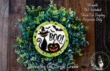 A Halloween-themed wreath made of green foliage and a black cat wearing a witch's hat, with a carved pumpkin and the word "BOO!" prominently displayed.