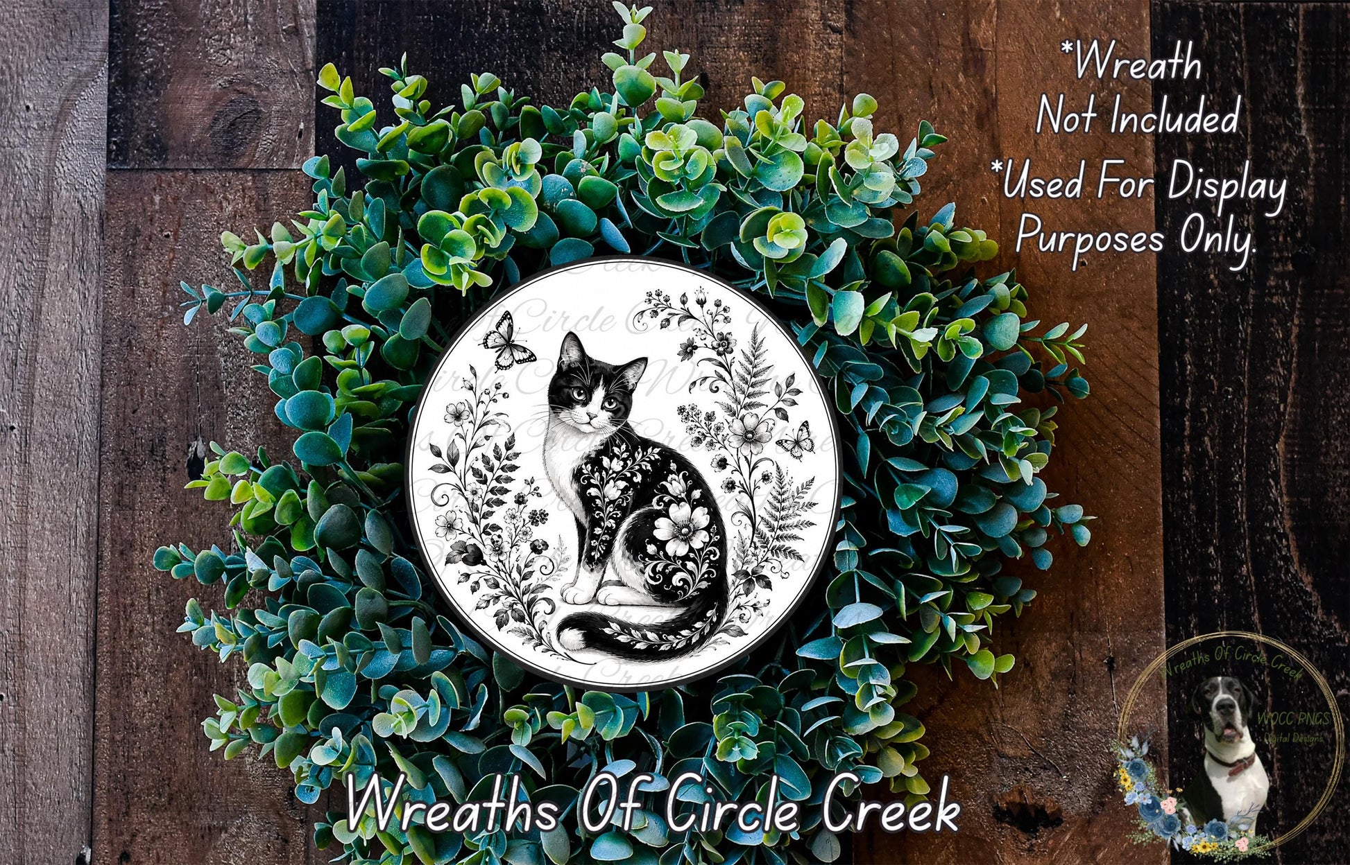 A wreath made of green plants and flowers, with a black and white illustration of a cat and a butterfly on it. The wreath is displayed on a wooden surface.