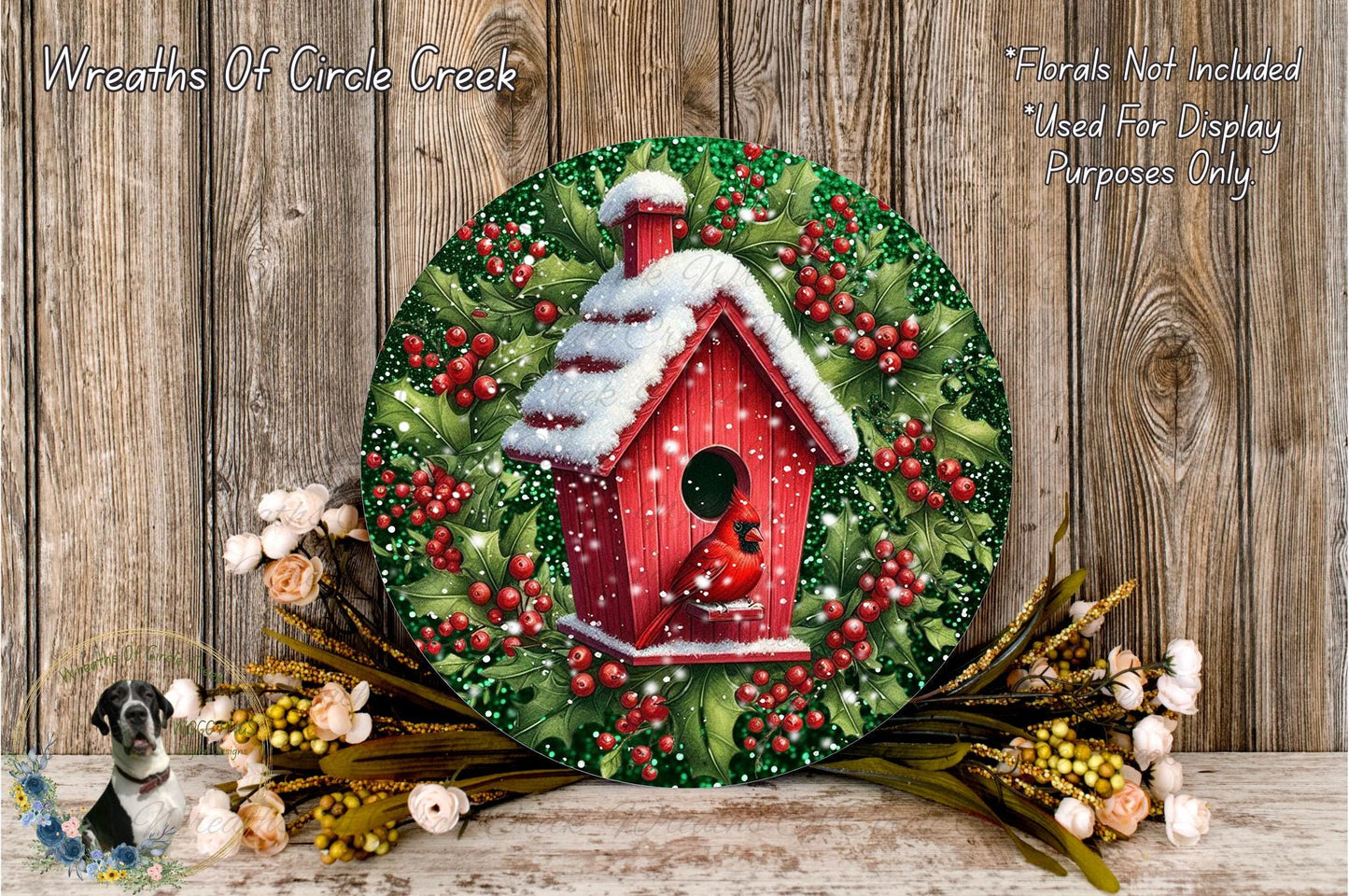 A round wooden wreath with a red birdhouse on top, surrounded by a mix of flowers and greenery, placed on a wooden surface.