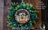 a Christmas wreath with a house and trees, surrounded by greenery. The wreath is displayed on a wooden surface, and there is a small dog in the bottom right corner of the image.
