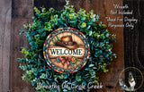 a wreath with a wooden sign that says "Welcome" surrounded by greenery. The wreath is displayed on a wooden surface, and there is a small dog in the bottom right corner of the image.