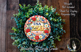 A wreath made of green leaves and flowers, with a circular plaque in the center displaying the text "USA 250 1776-2026" surrounded by red and white flowers.