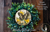 a wreath with a butterfly and yellow flowers, surrounded by green foliage. The wreath is displayed on a wooden surface, and there is a small dog in the bottom right corner of the image.