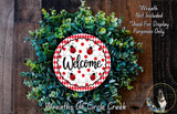 a wreath made of green leaves and a red and white welcome sign with ladybugs, placed on a wooden surface.