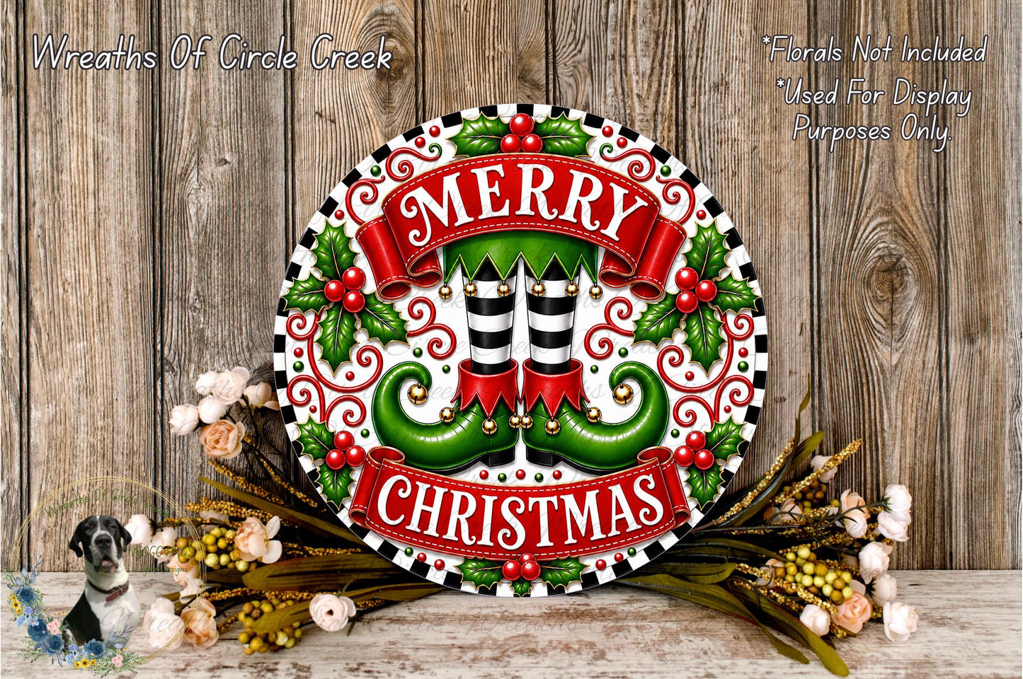 A festive Christmas wreath with the text "MERRY CHRISTMAS" displayed prominently. The wreath is adorned with green elf-like feet, red and white stripes, and red berries. The wreath is surrounded by a floral arrangement and placed on a wooden surface.