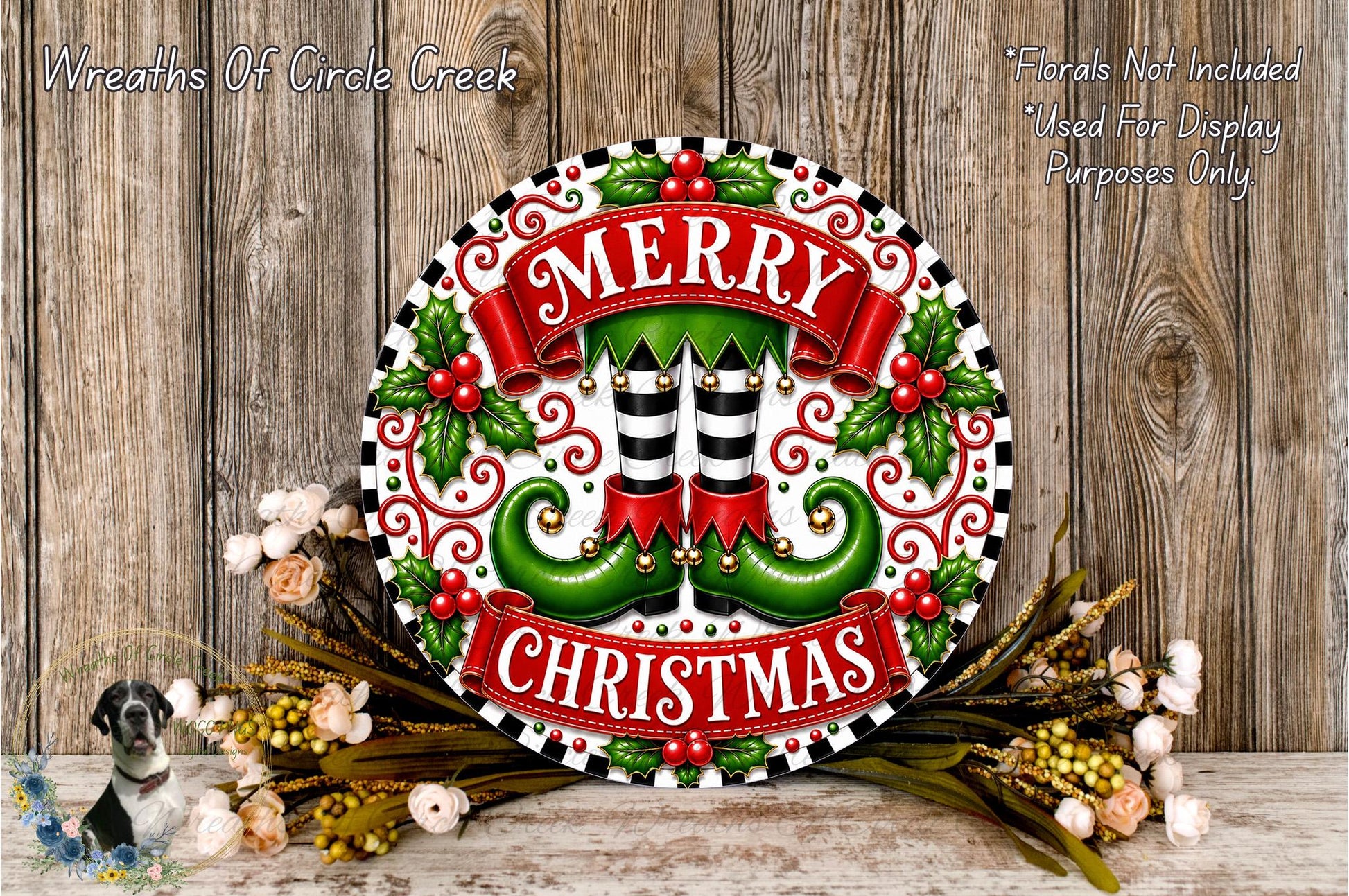 A festive Christmas wreath with the text "MERRY CHRISTMAS" displayed prominently. The wreath is adorned with green elf-like feet, red and white stripes, and red berries. The wreath is surrounded by a floral arrangement and placed on a wooden surface.