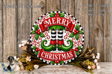 A festive Christmas wreath with the text "MERRY CHRISTMAS" displayed prominently. The wreath is adorned with green elf-like feet, red and white stripes, and red berries. The wreath is surrounded by a floral arrangement and placed on a wooden surface.