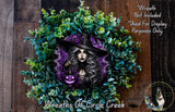 a wreath with a woman wearing a witch's hat and holding a lantern, surrounded by green foliage.