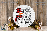 A white snowman sign with the text "Baby it's cold outside" and a snowman figure, surrounded by a wreath of flowers and a wooden background.