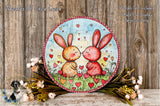 A round, decorative wreath with two rabbits in a field of flowers, surrounded by a floral arrangement and a wooden background.