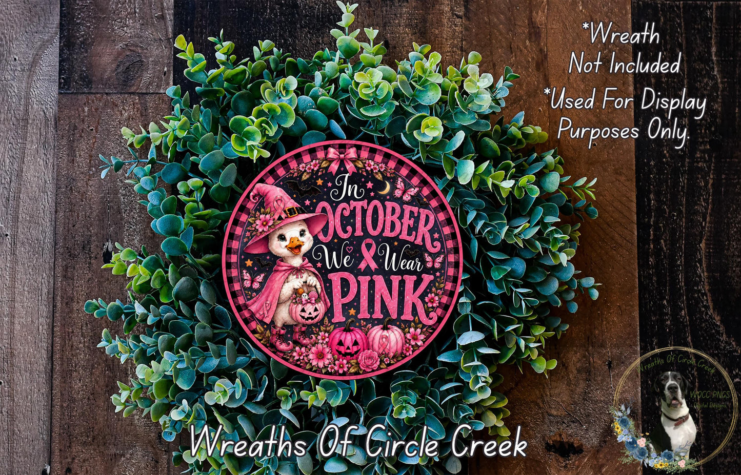A wreath made of green leaves and a pink circular sign with a witch and the text "In October We Wear Pink" is displayed on a wooden surface.