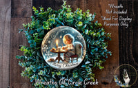 A wreath made of green leaves and a glass globe containing a child sitting at a table with a candle, surrounded by snowflakes, is displayed on a wooden surface.