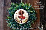 a wreath with a central image of a young girl wearing a red dress and a flower crown, surrounded by a lush green wreath.