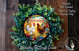 a wreath with a welcoming message, surrounded by greenery and flowers. The wreath is displayed on a wooden surface, and there is a small dog in the bottom right corner.