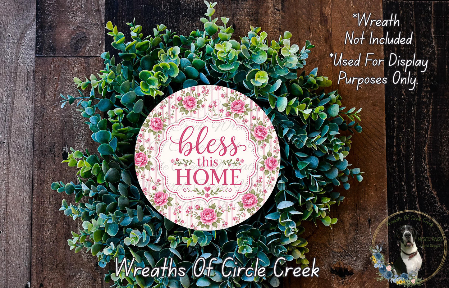 A wreath made of various green plants and flowers, with a pink and white sign that says "bless this home" in the center.
