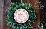 A wreath made of various green plants and flowers, with a pink and white sign that says "bless this home" in the center.