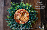 A wreath made of green leaves and flowers, with a pie and a jar of jam placed on top.