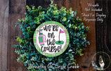 a wreath made of green foliage, with a circular sign in the center that reads "we're on the course!" The sign is surrounded by the wreath, and there is a small dog in the bottom right corner of the image.