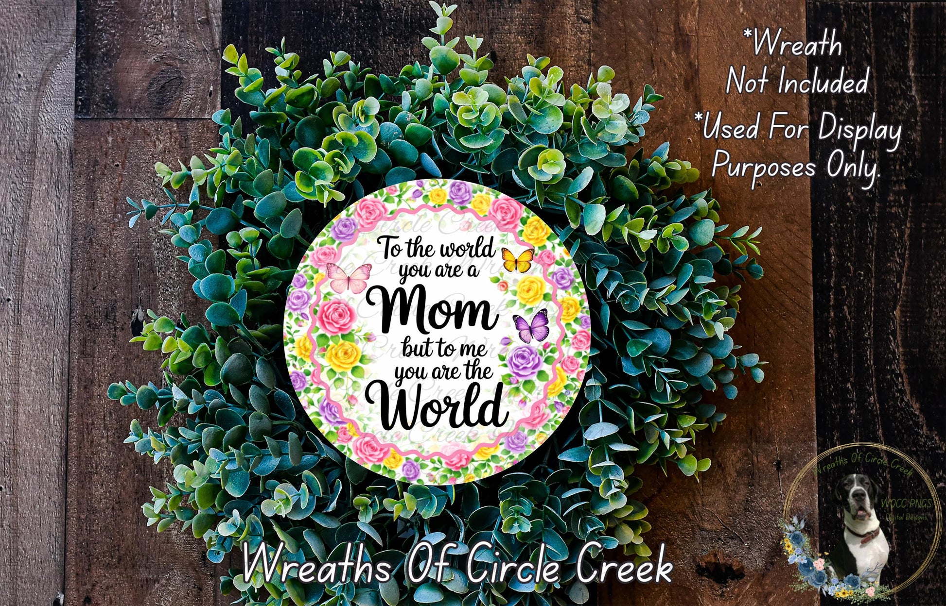 A wreath made of green leaves and flowers, with a circular sign in the center that reads "To the world you are a Mom but to me you are the World". The wreath is displayed on a wooden surface, and there is a small dog in the bottom right corner.