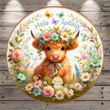 A round, decorative plate featuring a brown cow with horns, flowers, and butterflies surrounding it. The text "Hello Spring!" is written above the cow.