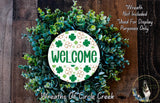 a wreath with the word "WELCOME" written on it, surrounded by various green and brown foliage. The wreath is placed on a wooden surface, and there is a small dog visible in the bottom right corner of the image.