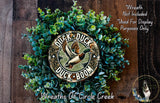 a wreath made of green foliage and a circular sign with the text "Duck, Duck, Duck" and a graphic of a duck in flight. The sign is surrounded by the green foliage, creating a decorative and nature-inspired arrangement.