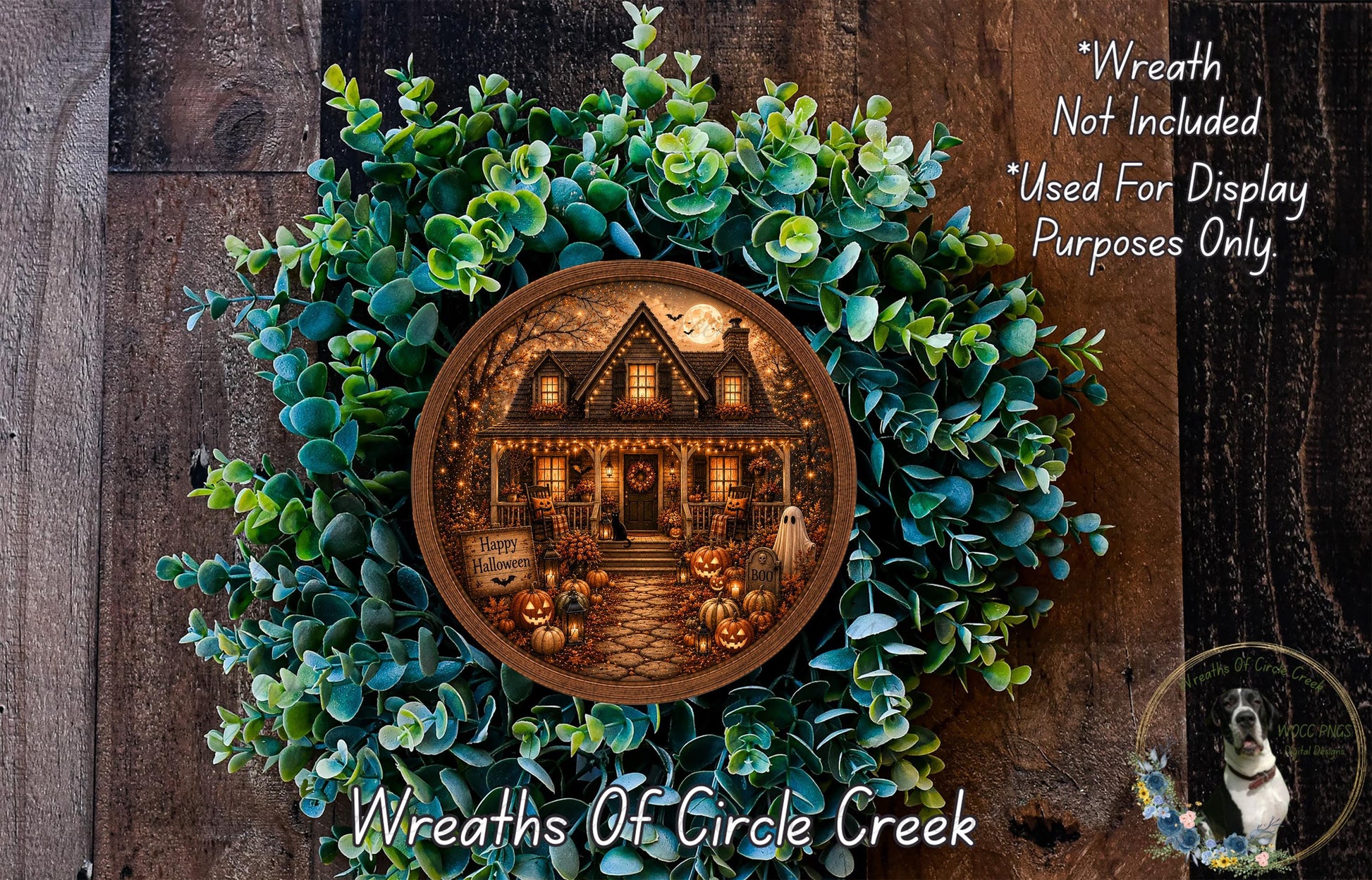 a wreath with a house scene inside, surrounded by greenery. The wreath is displayed on a wooden surface, and there is a small dog in the bottom right corner of the image.