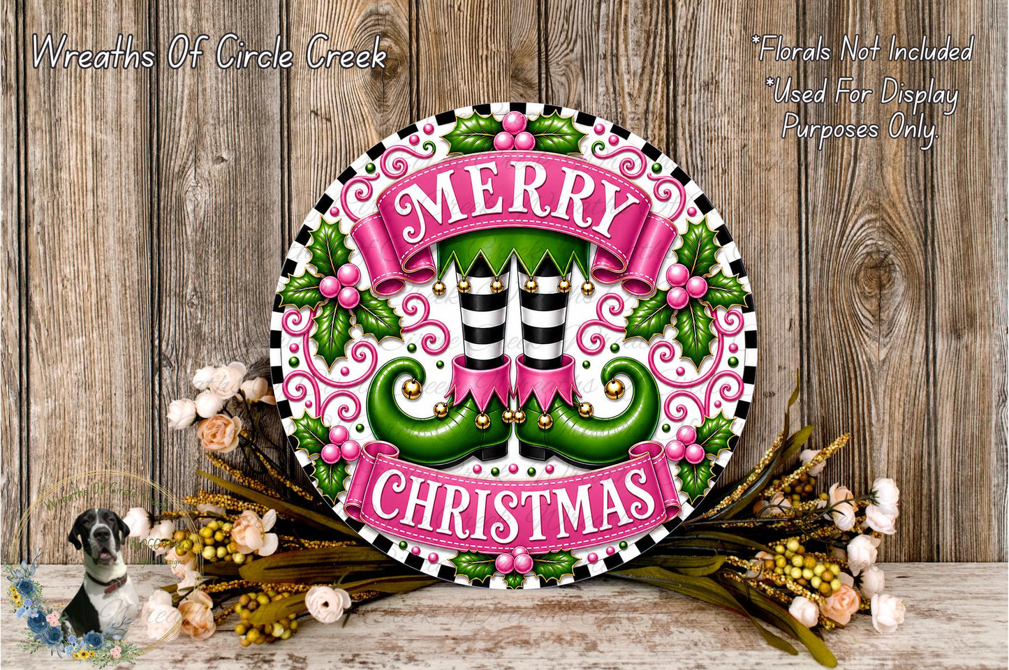 A festive Christmas wreath with the text "MERRY CHRISTMAS" displayed prominently. The wreath is adorned with green elf-like feet, a pink banner, and various holiday-themed elements such as flowers and leaves. The wreath is set against a rustic wooden background, creating a warm and inviting holiday atmosphere.