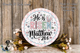 a decorative Easter-themed sign with the text &quot;He is risen Matthew 28:6&quot; surrounded by a floral arrangement and a wooden background.