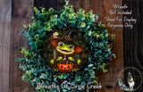 A Halloween-themed wreath featuring a frog wearing a witch's hat and holding a pumpkin, surrounded by green foliage.