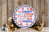 A decorative round sign with the text "She loves Jesus and America too" displayed on it, surrounded by a floral arrangement and a wooden background.