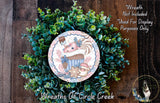A wreath made of green leaves and a decorative plate featuring a whimsical illustration of a cupcake and a cat.